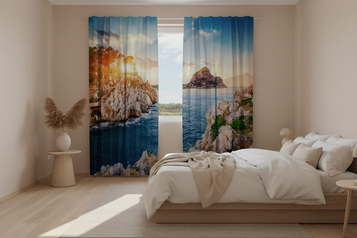 A bright bedroom interior with white walls and a wooden floor. Photo curtains are hung on the window, featuring a photorealistic depiction of a rocky Mediterranean landscape (the Sicilian coast). The left curtain panel shows steep, light-colored cliffs with green trees at the summit, behind which bright, golden sunlight shines. The right panel shows calm, dark blue sea water, a rocky shore in the foreground, and a large rocky island in the background.