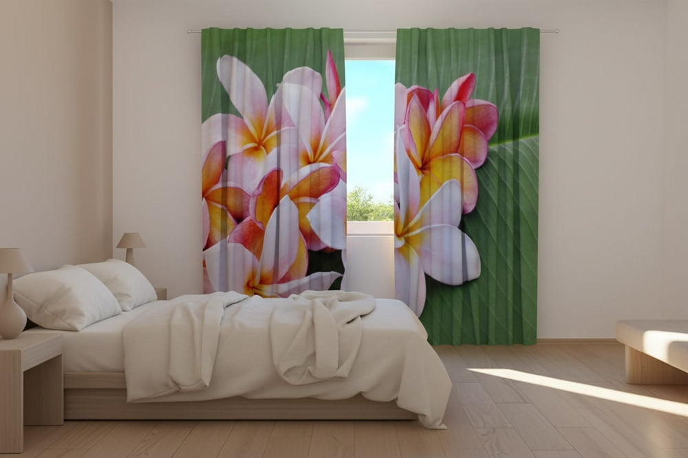 Gaišas un modernas guļamistabas interjers ar koka grīdu. Uz loga uzstādīti fotoaizkari, uz kuriem fotoreālistiski attēloti lieli balti, dzelteni un sārti plumerijas ziedi, kas izkārtoti uz lielas, zaļas tropu auga lapas.