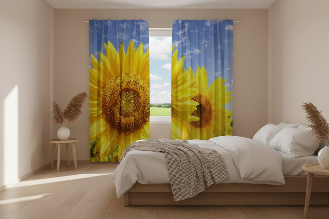 Gaišas un modernas guļamistabas interjers saules gaismā. Priekšplānā koka gulta ar baltu gultas veļu un pelēku pledu. Uz loga uzstādīti fotoaizkari, uz kuriem fotoreālistiski attēlotas liela izmēra spilgti dzeltenas saulespuķes uz zilu debesu un baltu mākoņu fona. Paverto aizkaru vidū ir logs ar skatu uz zaļu lauku. Telpu papildina koka naktsskapīši ar sauso augu dekorācijām vāzēs.