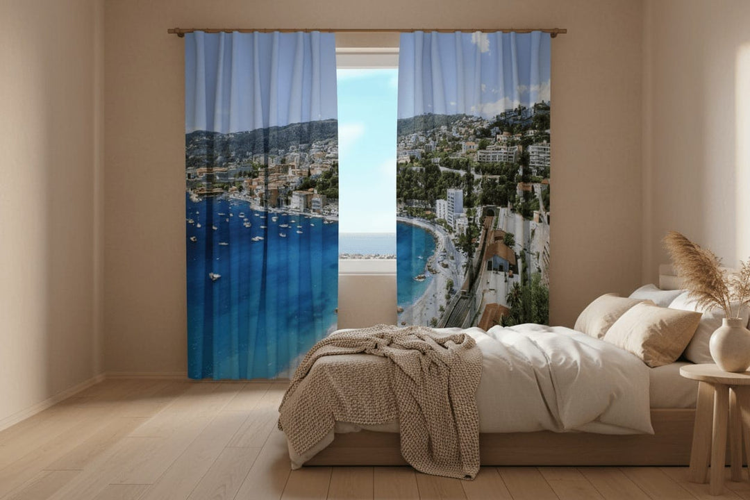A bright bedroom interior with a wooden floor and a light-colored bed. Photo curtains are hung on the window, featuring a photorealistic view of a picturesque Mediterranean (Greek) coastal town from a high vantage point.