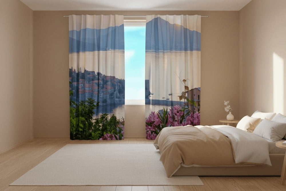 Bedroom interior with light-colored walls and a light-colored wooden floor. Photo curtains are hung on the window, depicting a Mediterranean coastal town in the light of sunrise or sunset in hazy pastel tones.