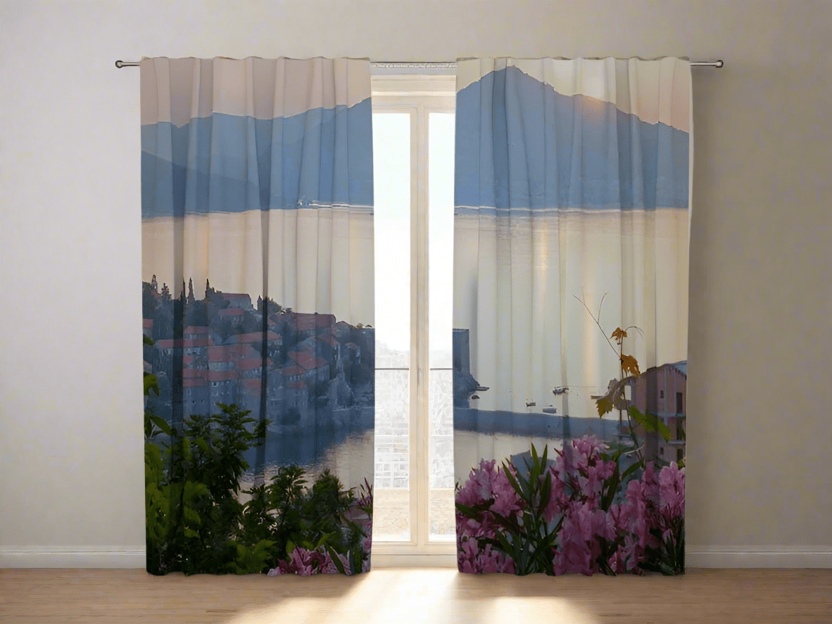 The room’s interior features white walls and a light-colored wooden floor bathed in natural sunlight. Photo curtains are hung on the window, depicting a Mediterranean coastal town in the light of sunrise or sunset in misty pastel tones. On the left side, the red tiled roofs of buildings at the foot of a hill and green trees are visible. The right panel shows calm sea water with boats and large mountain silhouettes in the background