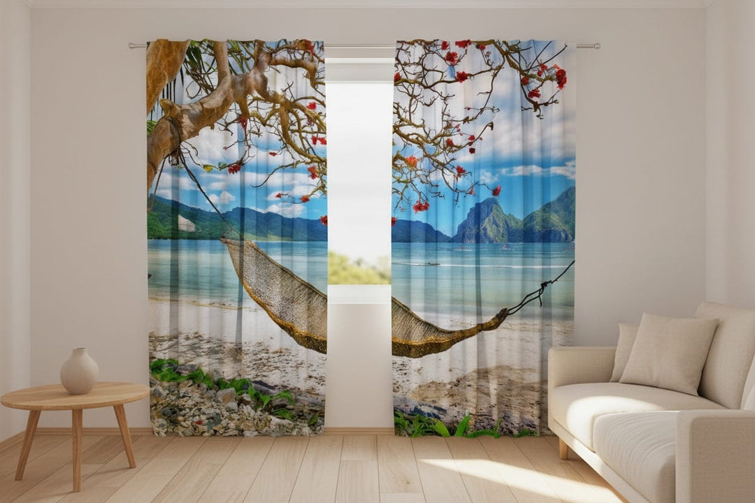 Living room interior with light-colored walls and a wooden floor. Photo curtains are hung on the window, featuring a photorealistic depiction of an exotic tropical beach. In the foreground, across the white sand and pebbles, a woven hammock is strung up, attached to a large, curved tree trunk with red flowers in its branches on the left side.