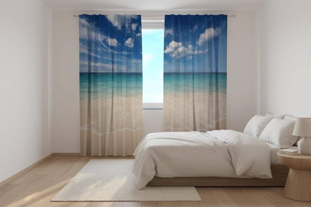 A bright and modern bedroom interior bathed in natural sunlight. To the right is a large bed in light tones. Photo curtains are hung on the window, featuring a photorealistic image of a tropical beach: light, golden sand with white sea foam, calm, turquoise ocean water, and a bright blue sky with white cumulus clouds.