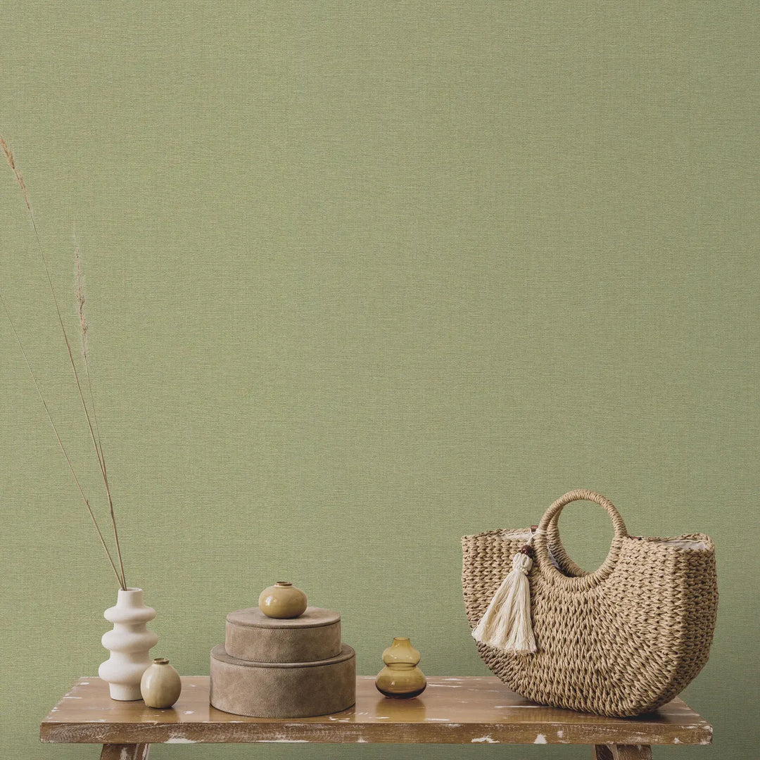 Olive green fabric-textured wallpaper in an interior with a wooden bench, decorative vases, and a woven bag