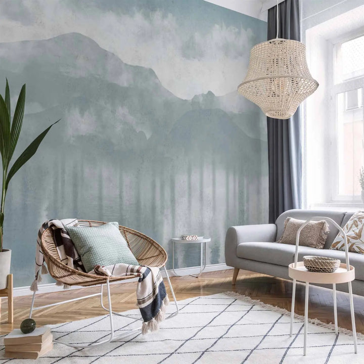 Photo wallpaper with a watercolor mountain landscape and reflection in water in light blue tones in a living room interior behind a wicker rocking chair, a grey sofa, and a natural wood parquet floor with a rug